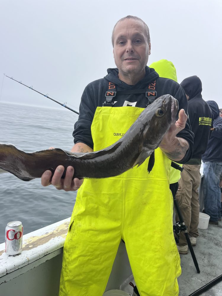Person in yellow rain gear holding a large fish on a boat, with a soda can and fishing rod visible.