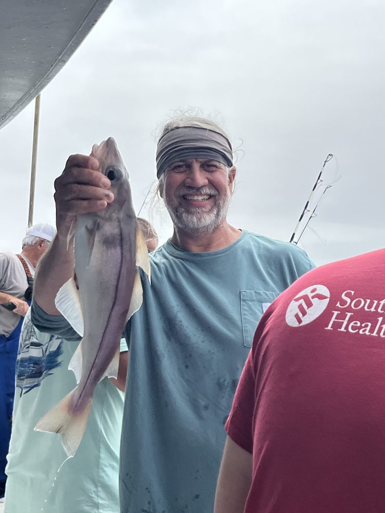 Man smiling and holding a fish on a boat with fishing rod in the background.