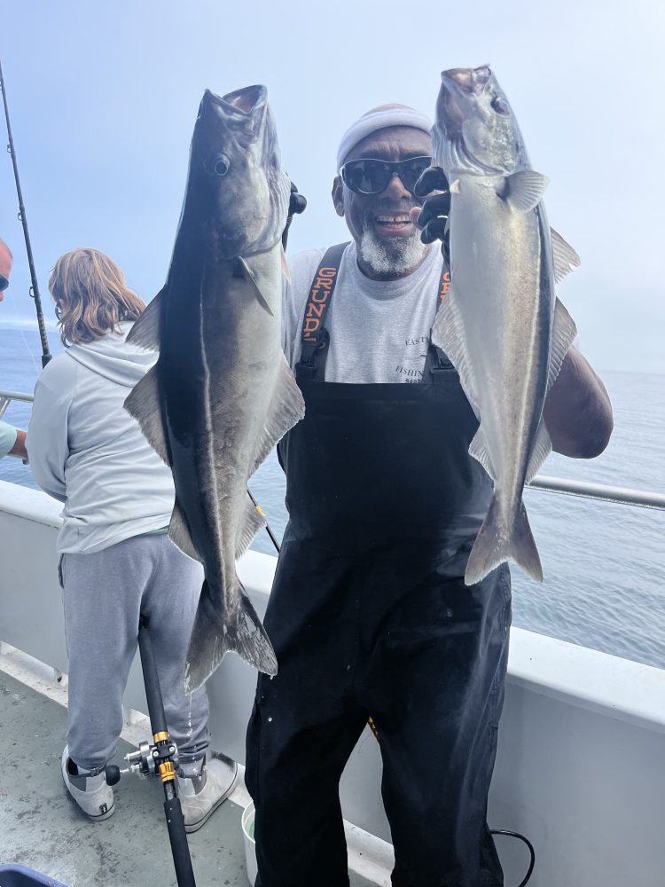 Person in black overalls holding two large fish on a boat.
