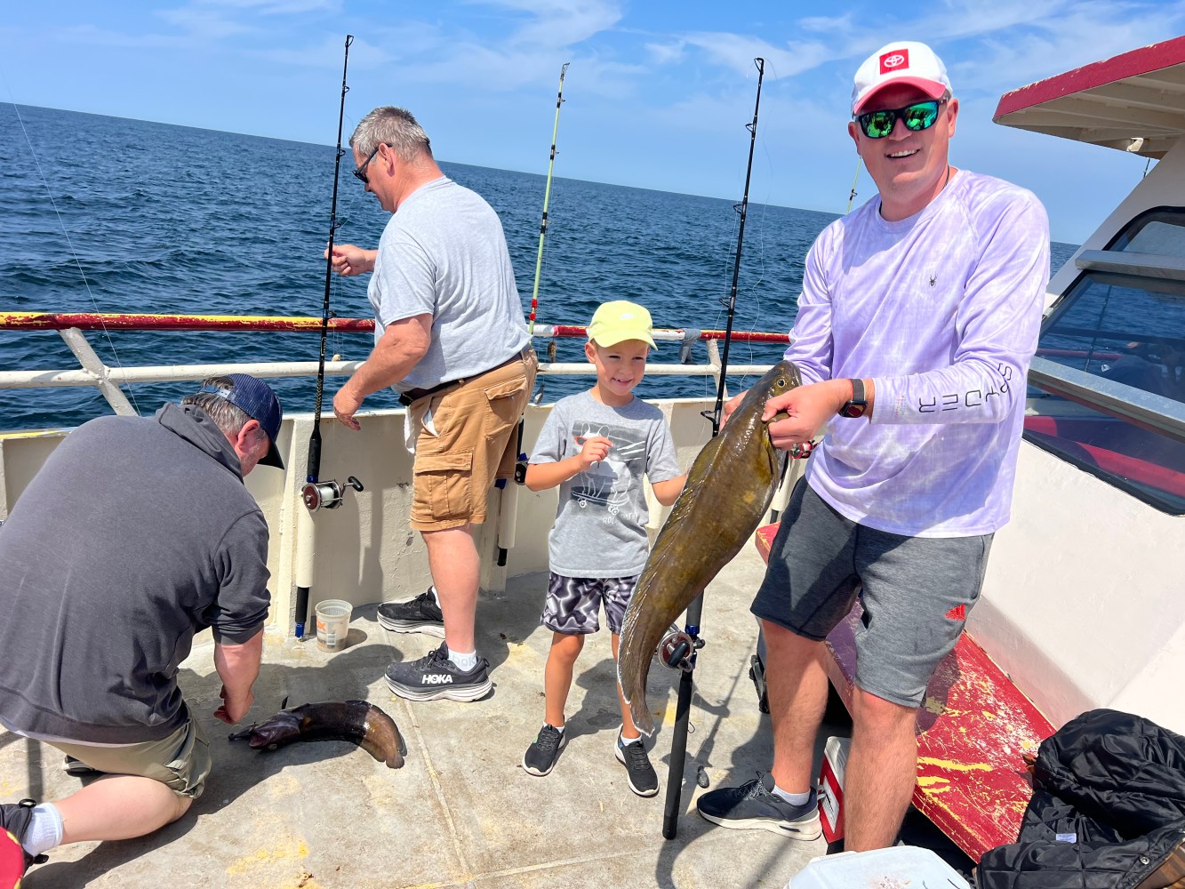 People on a boat with fishing gear; one holding a large fish and a child smiling beside him.