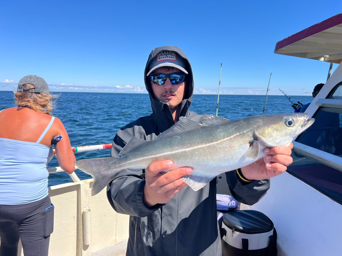 Person in sunglasses holds a fish on a boat with ocean in the background.