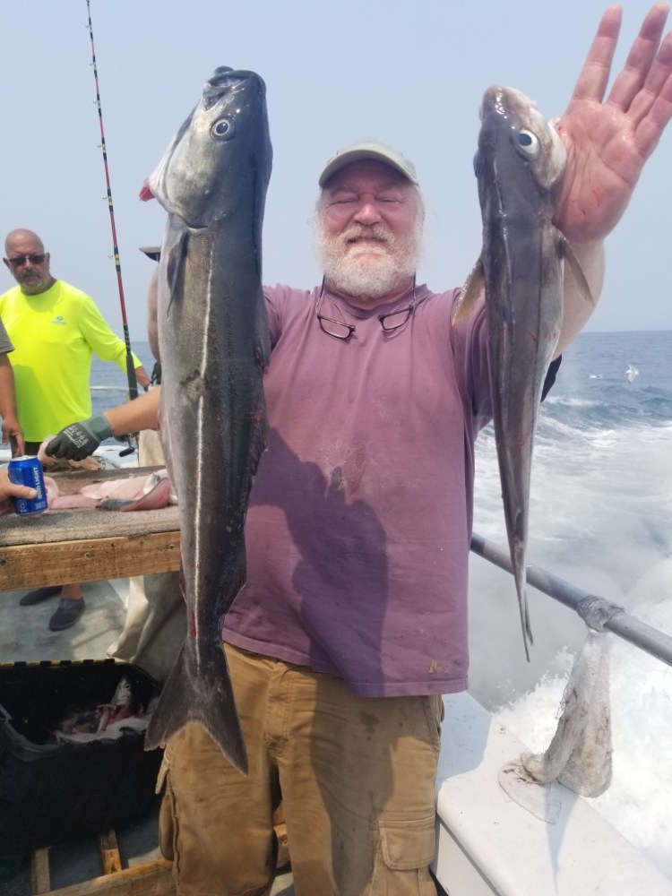 Man on boat holding two large fish, ocean and sky in background.