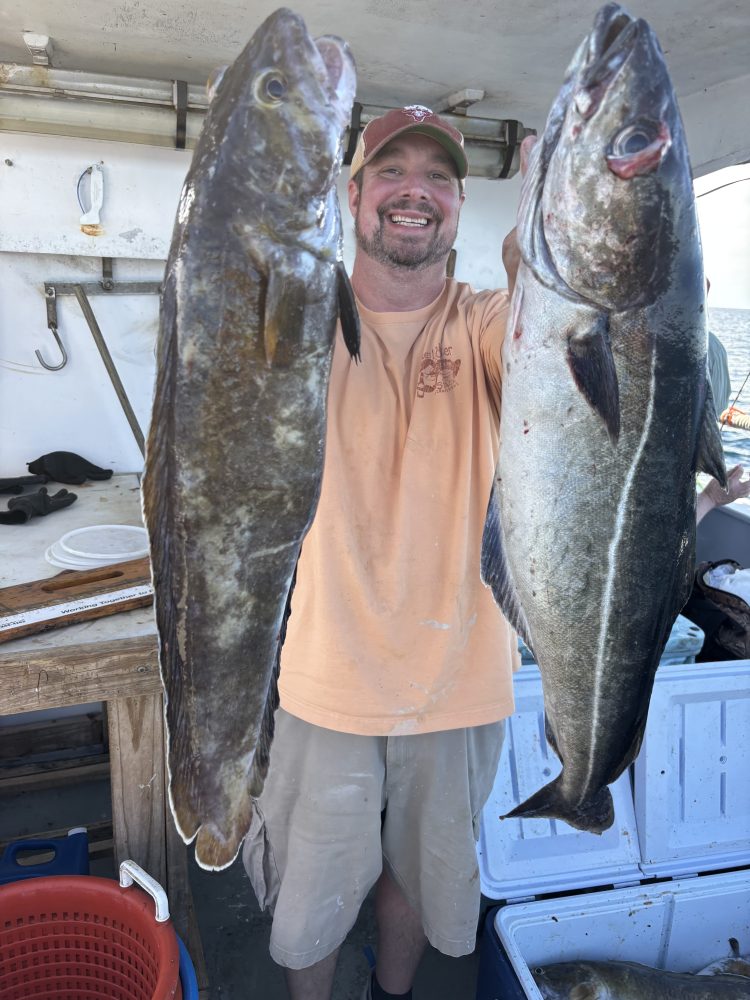 Man smiling, holding two large fish on a boat with a cooler and buckets nearby.