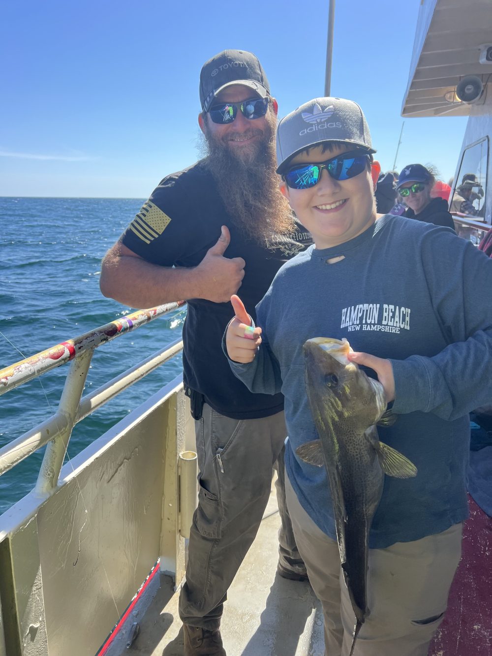 Two people on a boat holding a fish, both giving thumbs up, with ocean in the background.