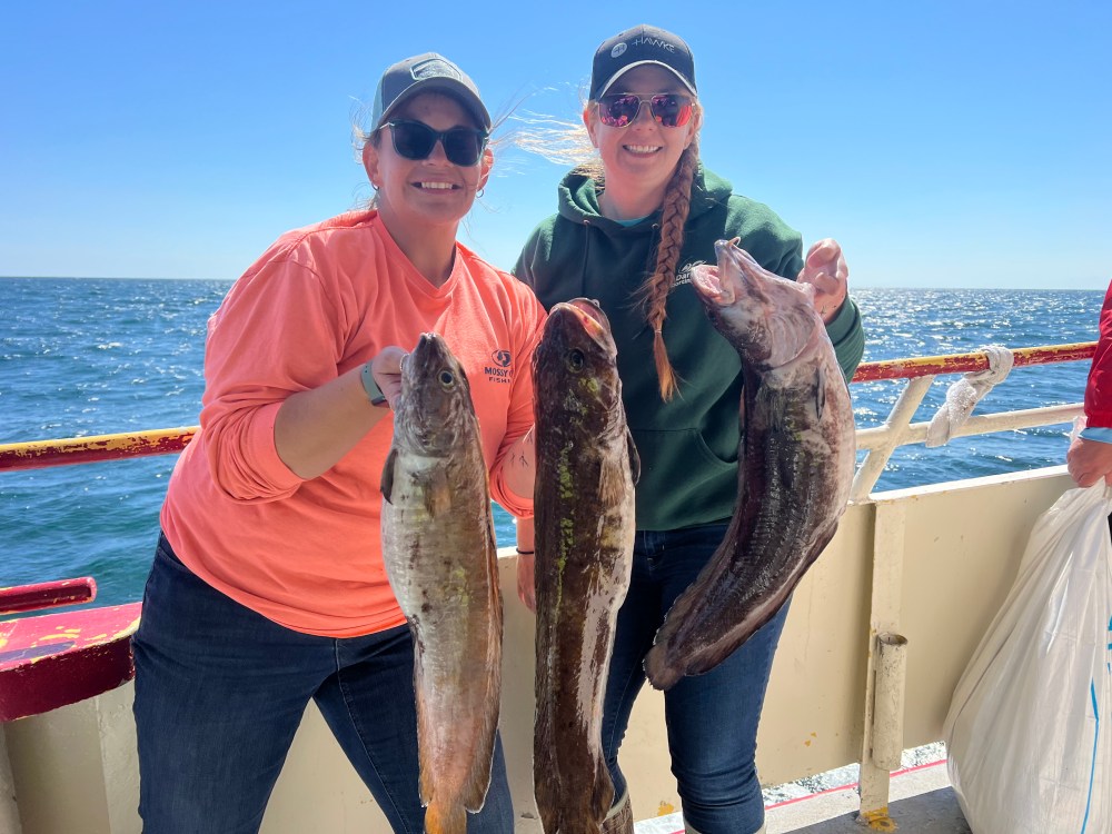 Two people on a boat holding large fish with the ocean in the background.