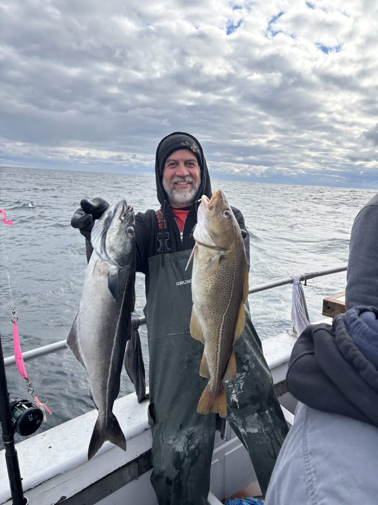 Person in a green jumpsuit holding two large fish on a boat with cloudy sky in the background.
