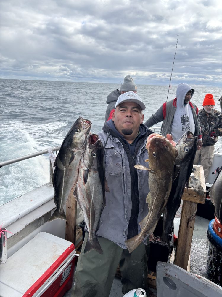Man on a boat holding five fish, ocean and overcast sky in background.