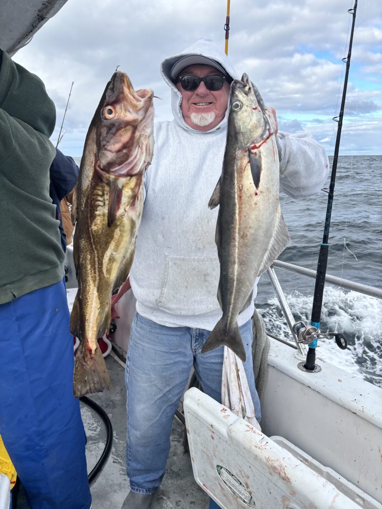 Man on boat holding two large fish, smiling, wearing a hoodie and sunglasses.