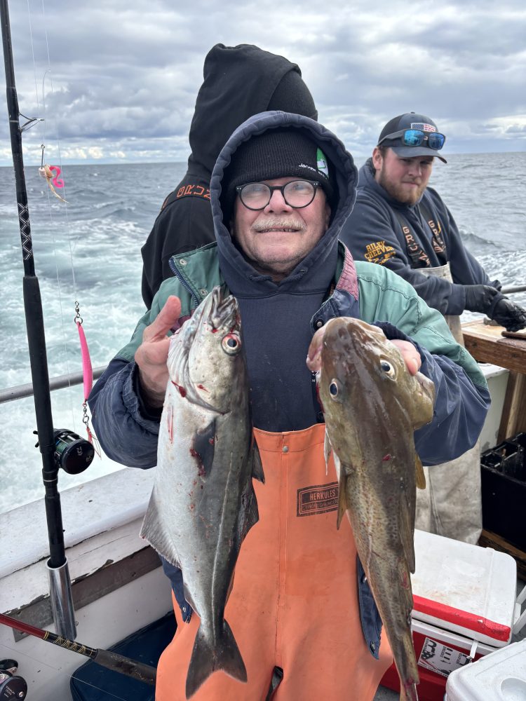 Person on a boat holding two fish with others in the background, overcast sky.