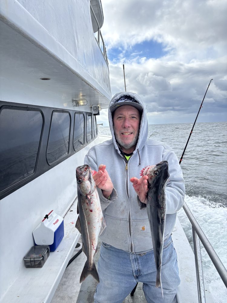 Person in hoodie on boat holding two fish with ocean and cloudy sky in background.