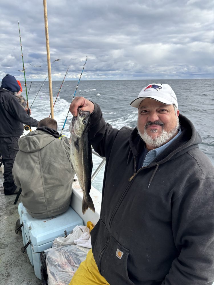Man on boat holding a fish, with the ocean in the background and fishing rods visible.