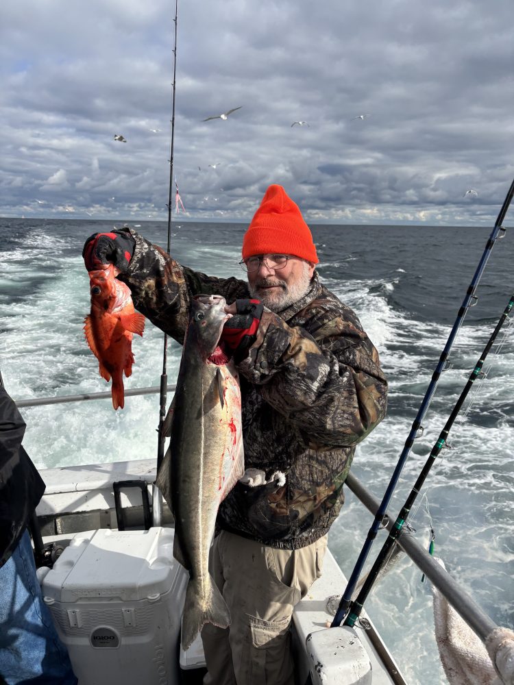 Man in red beanie holding two fish on a boat at sea with cloudy sky.