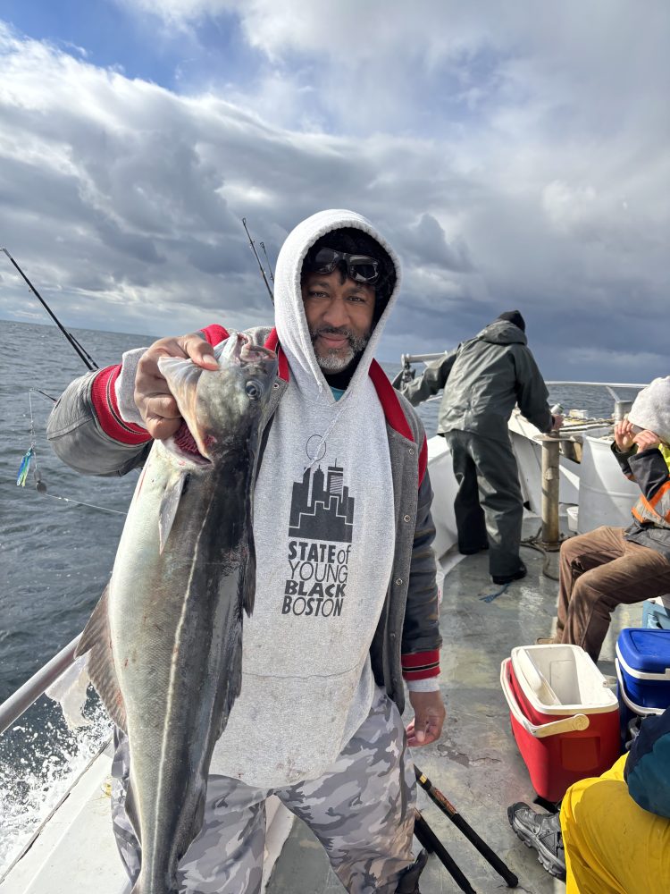 Person in a hoodie holding a fish on a boat with cloudy sky background.