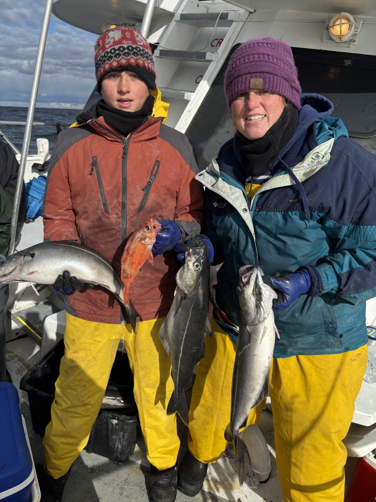 Two people in winter clothing holding fish on a boat deck.