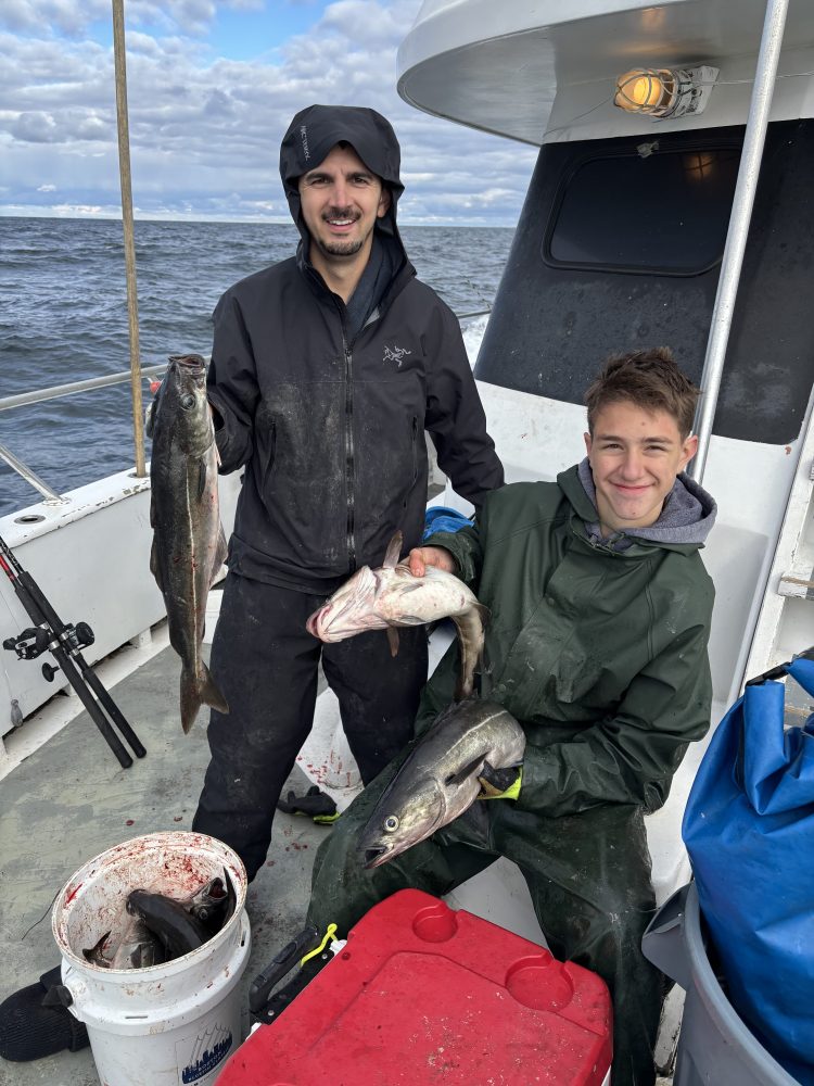 Two people on a boat holding freshly caught fish, with a bucket of fish on the deck.