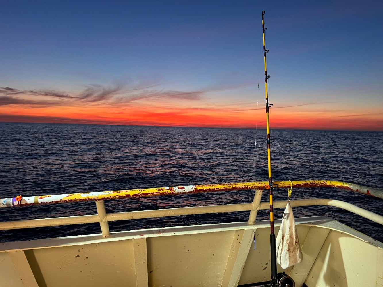 Fishing rod on a boat at sunset with a vivid orange sky and calm ocean.