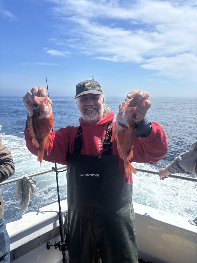 Man on a boat holding two fish with the ocean in the background.