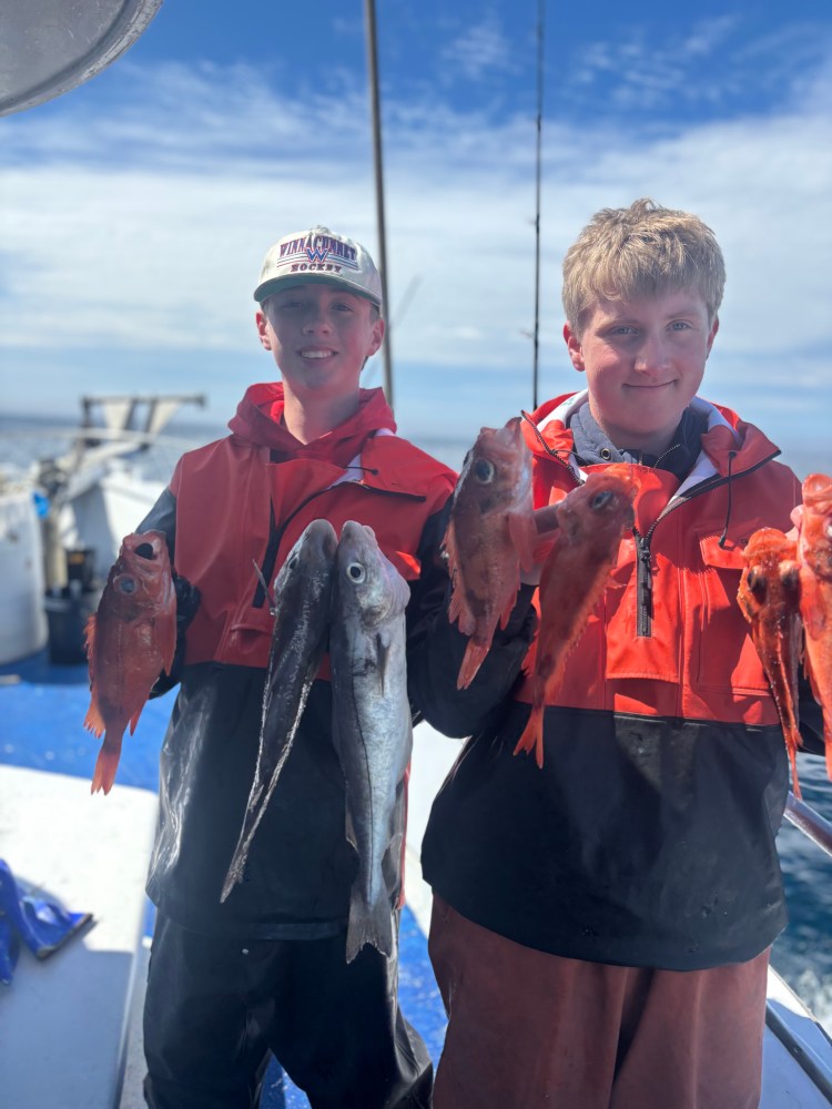 Two boys on a boat holding multiple fish, wearing red life jackets.