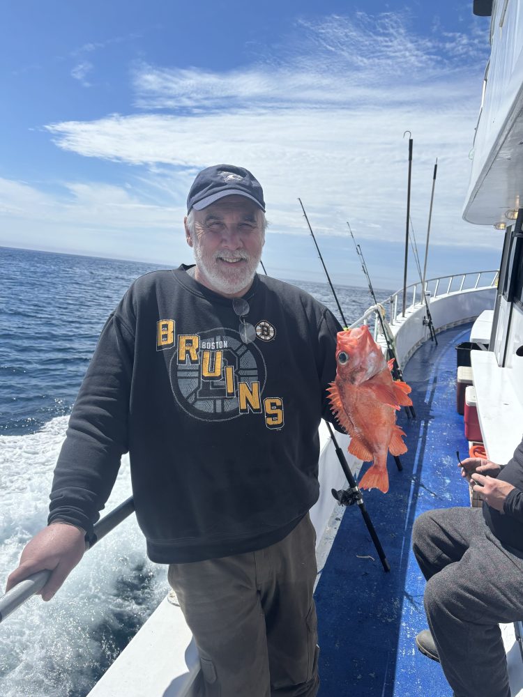 Man on boat holding an orange fish, wearing a dark sweatshirt, ocean in background.