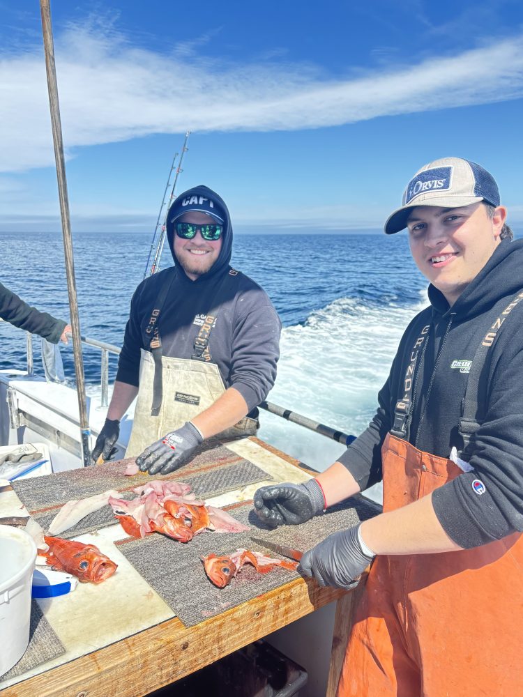 Two people on a boat filleting red fish on a table.