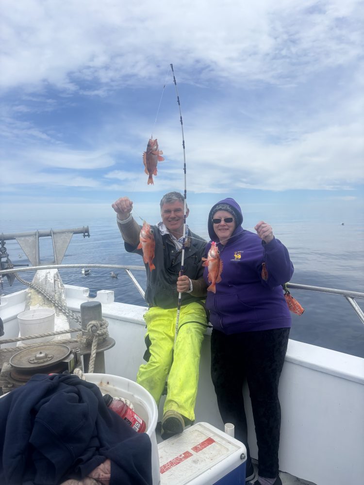 Two people on a boat holding fishing rods with fish, smiling.