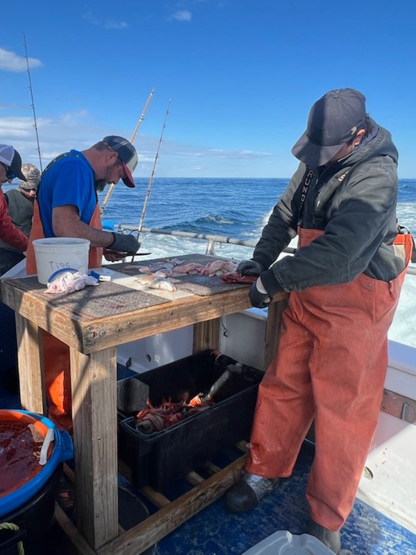 Two people in orange overalls clean fish on a boat with ocean in the background.