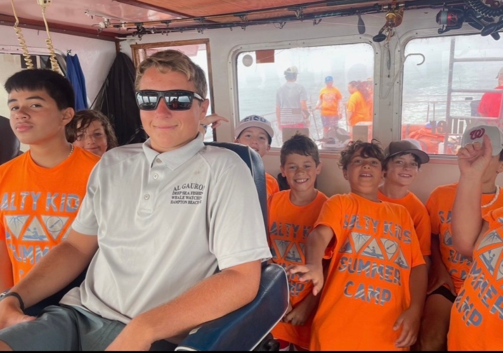 Group of kids in orange shirts on a boat posing with a man in a captain's chair wearing sunglasses.