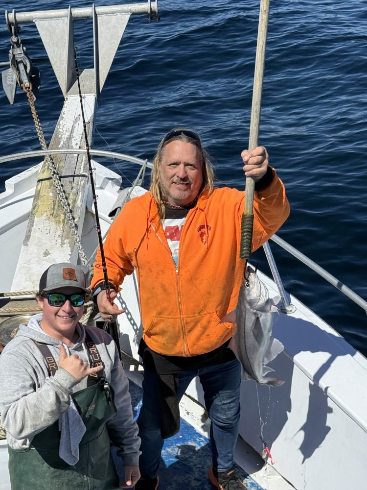Two people on a boat, one holding up a small fish with water in the background.