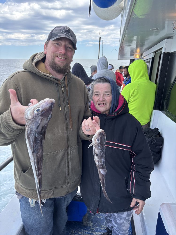 Two people on a boat holding freshly caught fish, smiling at the camera.