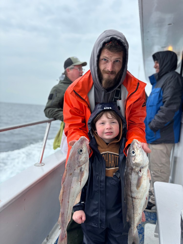 Two people on a boat holding large fish, with ocean in the background.