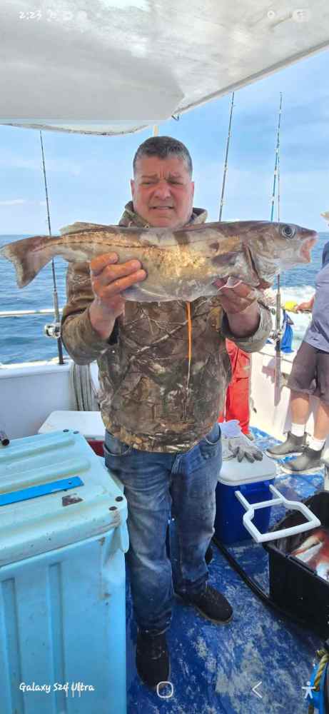 Man on boat holding a large fish, wearing camouflage jacket and jeans.