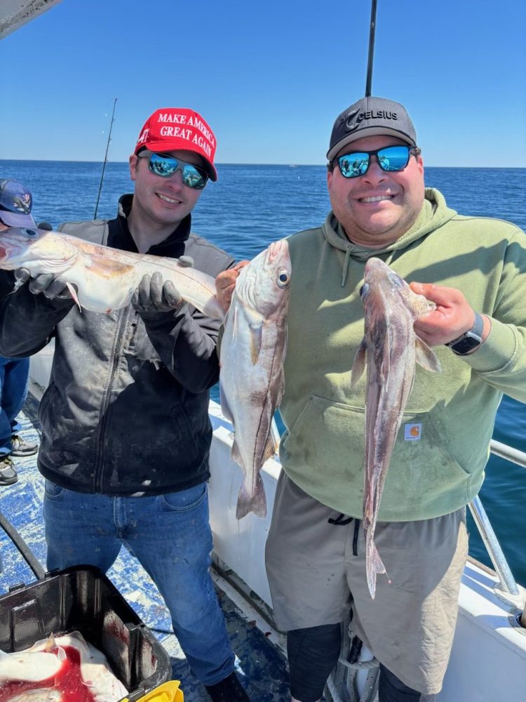 Two people on a boat holding large fish, with ocean in the background.