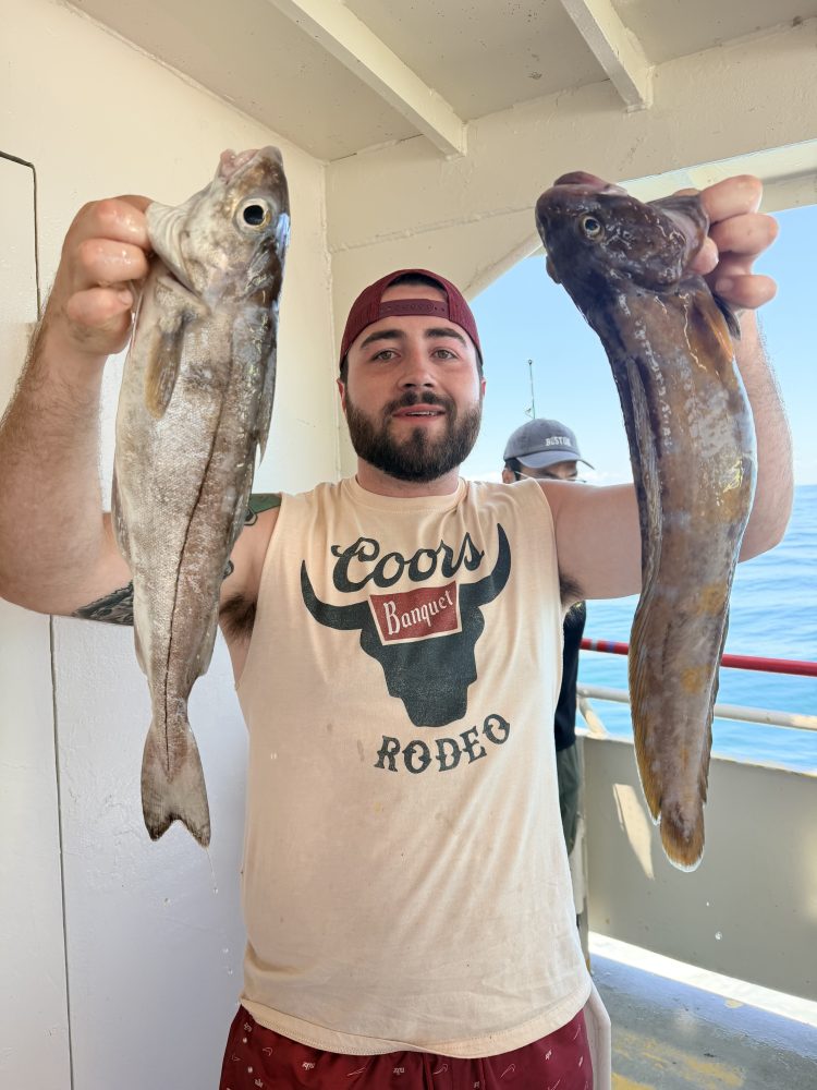 Man holding two fish on a boat, wearing red cap and Coors shirt.