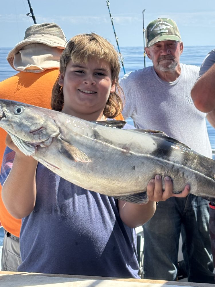 Boy smiling and holding a large fish on a boat with others around.