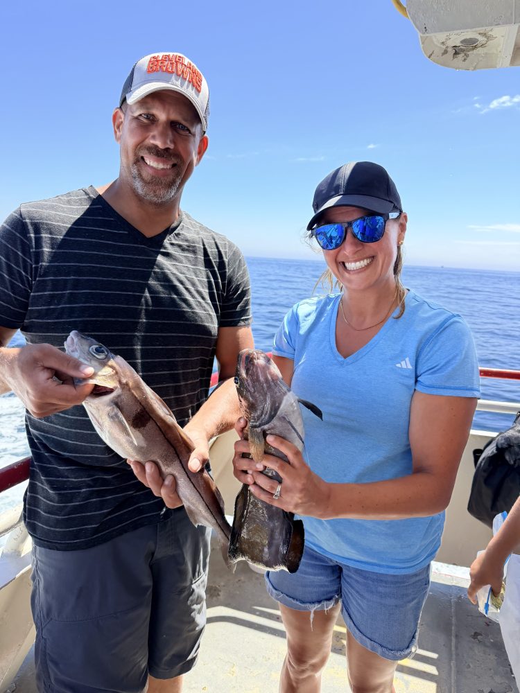Man and woman smiling, holding fish on a boat with ocean in background.