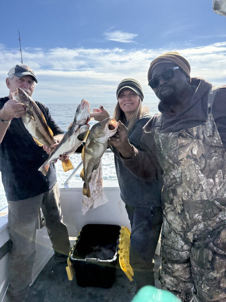 Three people on a boat holding fish with ocean in background.